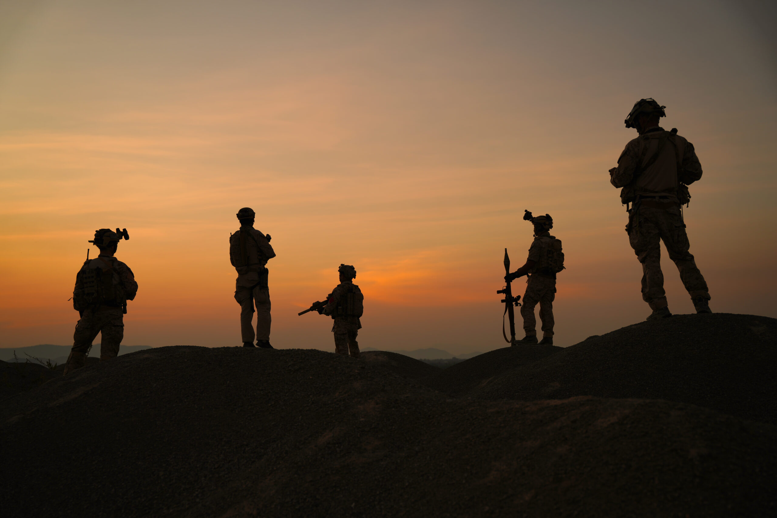 As the sun sets on the horizon, a group of soldiers can be seen silhouetted against the colorful sky. They are positioned on a rocky ridge, engaged in various activities, including checking equipment and maintaining communication. The atmosphere is tense yet focused as they prepare for an upcoming mission in the remote area. The fading light casts long shadows, emphasizing the determination and camaraderie among the troops as they strategize and await further instructions.